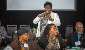 A woman in a white blazer is standing while speaking into a microphone and gesturing with her hands. She is addressing an audience during an event, with three attendees seated at a table in the foreground. The seated individuals, two women and one man, are attentively listening and wearing name tags. A large banner in the background displays the logo for N.C. A&T Cooperative Extension.