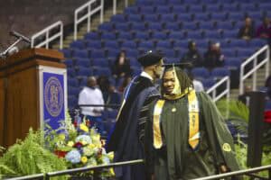 A graduate student wearing a black cap and gown with gold honor cords and an NC A&T sash walks across the stage during commencement. A university official in academic regalia stands in the background near a podium decorated with the NC A&T seal.