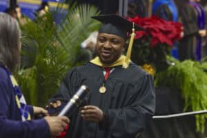 A graduate student in cap and gown receives a diploma during commencement. The student, smiling, wears a black graduation cap and gown with a yellow hood, symbolizing their academic achievement.