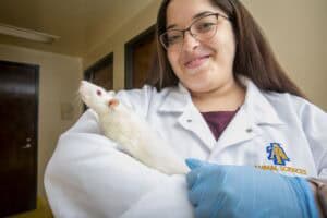 A student wearing a white lab coat with the NC A&T Animal Sciences logo holds a white laboratory rat. She smiles warmly while wearing blue protective gloves, standing in a research facility hallway.