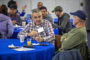 Two men are seated at a round table with a blue tablecloth, engaged in conversation. One man, wearing a plaid shirt and a name tag, is speaking while gesturing with his hand. The other man, wearing a green sweater and a blue baseball cap, is listening. Other attendees are visible in the background, seated at similar tables.
