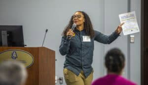 A woman with curly hair and glasses is speaking into a microphone while holding a printed program for the World Soil Day Celebration. She is wearing a dark green shirt with a name tag and is gesturing with her other hand. A podium with the North Carolina Agricultural and Technical State University logo and a computer monitor are visible in the background.