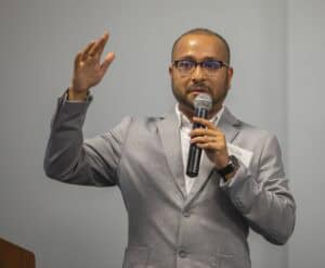 A man wearing glasses and a gray suit is holding a microphone while raising his other hand as he speaks. He has a name tag pinned to his suit jacket. The background features a neutral-colored wall.