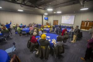 A large group of people is seated at round tables with blue tablecloths in a conference room. A speaker at a podium in front of a screen is addressing the audience. The screen displays the text "World Soil Day Celebration." Attendees are listening, some taking notes, while a person with a camera is recording the event. The room has gray walls, overhead lights, and a carpeted floor.