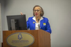 A woman with short curly hair and glasses is speaking at a podium. She is wearing a bright blue blazer, white blouse, and a name tag, gesturing with one hand while standing behind a microphone. A large monitor and the North Carolina Agricultural and Technical State University logo are visible on the podium.