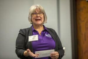 A woman with glasses and short gray hair is speaking while gesturing with her hand. She is wearing a purple polo shirt with a logo, a black jacket, and a name tag that reads "Kristina Hare." She is holding a piece of paper. The background features a wooden door and a gray wall.