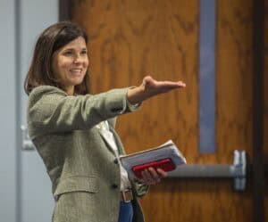 A woman in a blazer and white blouse is smiling while gesturing with her hand during a presentation. She holds a notebook and a red smartphone in her other hand. A wooden door with a narrow vertical window is visible in the background.