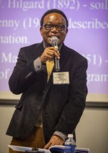 A man identified as Dr. Uzo, wearing glasses and a black blazer, holds a microphone while moderating a panel discussion at the World Soil Day Celebration.