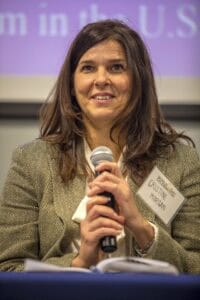 A woman identified as Cristine Morgan, wearing a herringbone blazer and holding a microphone, smiles while speaking during a panel discussion at the World Soil Day Celebration.