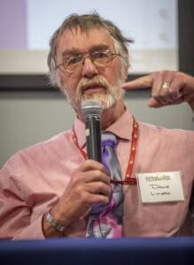 A man identified as David Lindbo, wearing glasses, a pink shirt, and a purple tie, speaks into a microphone and gestures with his hand during a panel discussion.