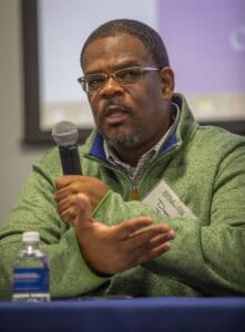 A man identified as Dedrick Davis, wearing glasses and a green sweater, speaks into a microphone while gesturing with his hand during a panel discussion.
