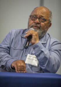 A man with glasses, identified as Millard Locklear, speaks into a microphone while seated at a table during a panel discussion at the World Soil Day Celebration.