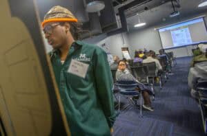 A student wearing a green MANRRS shirt and name tag reading "Miles Lee" examines a poster session while attendees sit in rows watching a presentation on a large screen in the background.