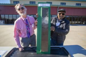 Two men hold up a green-framed soil profile labeled “Leon” during a soil demonstration outside a red building. One man wears a pink shirt and tie, and the other wears a black jacket and hat.