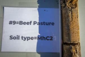A close-up of a soil sample displayed alongside a printed label reading “#9 = Beef Pasture, Soil type = MhC2” during a soil demonstration.