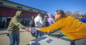 A woman in a mustard-yellow sweater hands a document to a man in a green hoodie during an outdoor soil health event, with participants and a red building in the background.