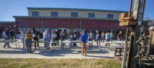 A large group of people observes a soil quality demonstration led by a person in a blue jacket outside a red building. A tall soil testing machine is set up nearby.