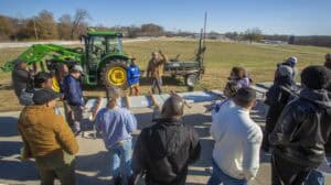 A group of people gathers around tables to observe a soil quality demonstration conducted by two individuals near a green tractor and soil testing equipment in an open field.