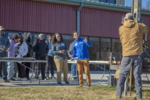 A group of people observes a soil test demonstration outside a red building. A man in a blue jacket gives a thumbs-up while signaling another person operating soil testing equipment.