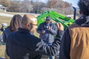 A soil scientist, wearing a black jacket and hat, speaks into a microphone while demonstrating soil quality at an outdoor event, with a green tractor in the background and attendees in the foreground.