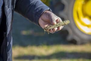 Close-up of a person’s hand holding a soil sample with roots attached during an outdoor soil demonstration, with a yellow tractor tire visible in the background.