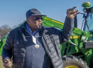 A soil scientist wearing a black cap, sunglasses, and a black puffer jacket holds up a small soil sieve while speaking during a soil quality demonstration. A green tractor is visible in the background, highlighting an outdoor farm setting.