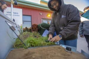 Two individuals closely examine a soil display during a rainfall simulation demonstration. A woman in the foreground, wearing a brown Carhartt jacket and glasses, adjusts or examines the plants in a soil tray. Another individual with red braids and a green jacket leans in to observe the soil demonstration. The USDA NRCS Soil Health Division sign is visible in the background, alongside a red corrugated metal building.