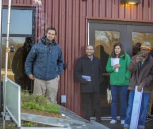 Four people are gathered outside a red corrugated metal building, watching a water demonstration. The man on the left wears a blue jacket and beige pants, smiling while observing the water feature. Next to him, a man in a black coat holds papers, smiling. A woman in a green jacket holding a notebook stands beside him. On the far right, a man in a brown jacket and orange hat holds a phone, seemingly recording or photographing the scene. In the foreground, a sloped metal tray displays plants and soil.