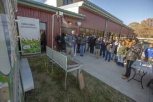 A man operates a rainfall simulator during an outdoor demonstration, spraying water over soil samples in front of an audience. The display highlights how different soil management practices affect runoff and infiltration.