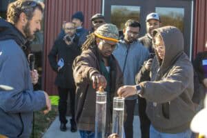 Two undergraduate students assist a soil specialist in a rainfall simulation demonstration, placing soil samples into tall water-filled containers to observe their effects. A small audience watches the demonstration in the background.