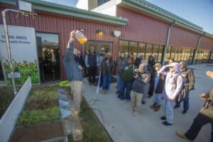 A man demonstrates a rainfall simulator to a group of attendees outdoors, holding up a container of water runoff to illustrate soil management effects. Several soil samples with varying vegetation are displayed to compare infiltration and runoff.