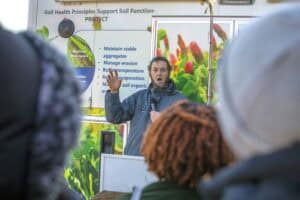 A man gives a soil health presentation in front of an educational display that highlights principles like managing erosion, buffering temperature, and maximizing soil organic matter. Attendees listen to the speaker outdoors in winter clothing.