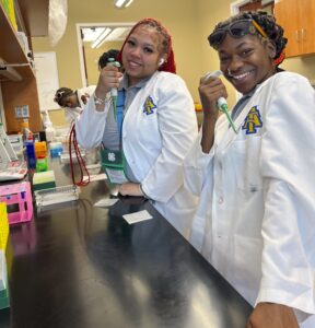 Two students in white lab coats, featuring the logo of North Carolina A&T State University, smile while holding pipettes in a laboratory. Both students stand at a lab bench with colorful lab equipment, including racks and trays, in front of them. In the background, other students in lab coats are engaged in activities, and a whiteboard with diagrams is visible on the wall.