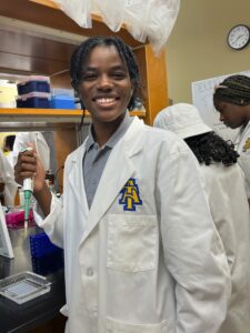 A smiling student in a white lab coat, featuring the logo of North Carolina A&T State University, holds a pipette in a laboratory. The student stands near a lab bench with various equipment and containers visible. Behind them, other students wearing lab coats and hats are engaged in laboratory work. A clock and a whiteboard with handwritten notes are visible in the background.
