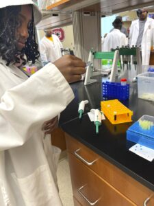 A young scientist in a white lab coat and a white bucket hat works in a laboratory. They are holding a small vial, focusing on its contents. The lab bench in front of them contains pipettes, a yellow tube rack, a blue container with test tubes, and other lab equipment. In the background, other individuals in lab coats are visible, engaging in laboratory activities.