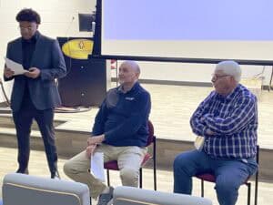 Three men are in a classroom setting. A young man wearing a dark suit stands to the left holding papers, appearing to read or present. Two older men sit on chairs to the right; one wears a dark pullover and beige pants while the other wears a plaid shirt and jeans. A projector screen and lectern are visible in the background.