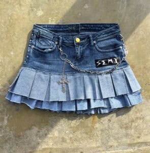 Denim skirt with pleated hem, chain detail, cross pendant, and a "SEMT" label, laid on a textured surface.