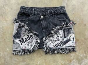 A pair of black denim shorts with colorful fringe and patchwork decorations featuring text and patterns.