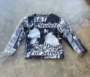 A black and white sweater featuring newspaper-style graphics and text about A&T students and sit-ins.
