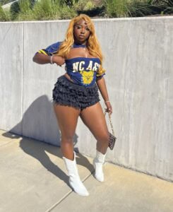 A woman poses confidently in a blue NC A&T shirt and black ruffled shorts, wearing white boots and holding a purse.