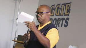 A middle-aged man with glasses and a beard, wearing a yellow polo shirt and a navy blue vest, speaks into a microphone while holding a piece of paper. He appears to be delivering a speech or presentation. Behind him, a wall displays the words 