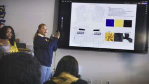 A woman in a navy blue sweater and white collared shirt presents a design concept on a large screen in a classroom or meeting room. She holds a microphone in one hand and gestures toward the screen with the other. The screen displays a glove design labeled "Aggie Esports," including material specifications and color swatches in blue, yellow, black, and white. Several audience members, some wearing matching yellow and navy blue attire, are seated and listening. Another woman in a yellow shirt stands nearby, observing the presentation. The room has a professional yet informal setting with power outlets visible on the wall under the screen.