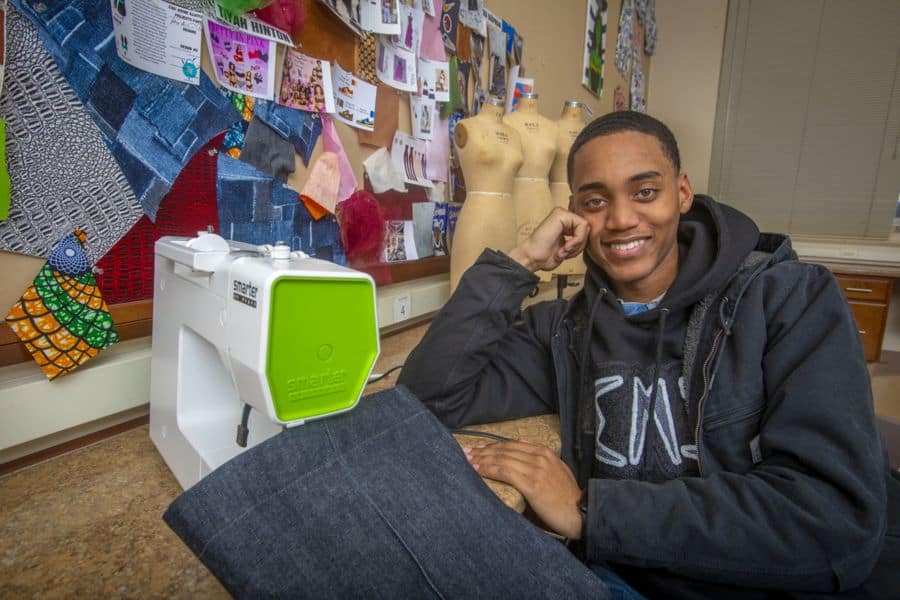 A young man with short black hair and a warm smile sits at a sewing workstation, leaning on his hand. He wears a black hoodie with a graphic design and a dark jacket. In front of him, a white and green sewing machine is placed on the table, with a piece of dark denim fabric draped over it. Behind him, a bulletin board is covered with fabric swatches, sketches, and pinned images. Two dress forms stand in the background, adding to the creative workspace atmosphere.