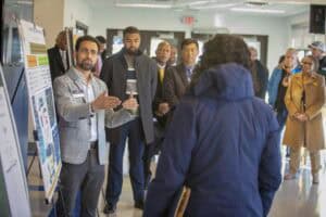 A man with short dark hair and a beard, wearing a gray blazer and a name badge, gestures while speaking to a woman in a navy blue coat, who listens attentively. He stands next to a large poster board with charts and graphics. Behind them, a diverse group of people, including men and women in business attire and coats, observe the presentation. The setting is an indoor hallway with large glass doors in the background allowing natural light to enter.
