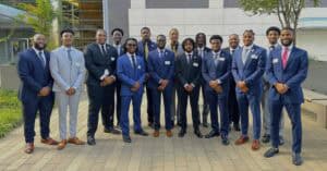 A group of 17 young African American men pose outdoors in front of a modern building with large glass windows and a white facade. They are dressed in formal business attire, wearing suits in shades of navy, black, and gray, paired with dress shirts and ties. Some of them have name tags pinned to their jackets. The group stands on a paved courtyard with greenery and a tree nearby, smiling confidently at the camera.