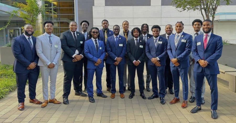 A group of 17 young African American men pose outdoors in front of a modern building with large glass windows and a white facade. They are dressed in formal business attire, wearing suits in shades of navy, black, and gray, paired with dress shirts and ties. Some of them have name tags pinned to their jackets. The group stands on a paved courtyard with greenery and a tree nearby, smiling confidently at the camera.