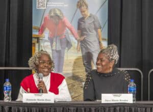 Two older African American women sit at a panel discussion table, smiling at each other. The woman on the left, Jeannette Martin Horn, wears a red and white sweater and holds a microphone while speaking. The woman on the right, Joyce Martin Bowden, wears a black sweater adorned with gold embellishments and listens attentively. Name cards in front of them display their names. Two bottles of water are placed on the table. Behind them, a large poster features an image of two women walking inside a greenhouse, with text promoting the Cooperative Extension at N.C. A&T and Small Farms Week.