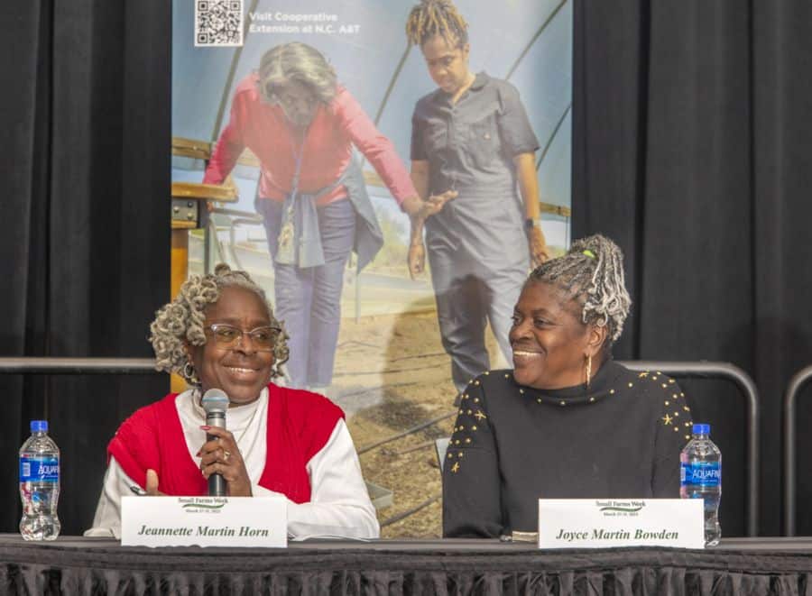 Two older African American women sit at a panel discussion table, smiling at each other. The woman on the left, Jeannette Martin Horn, wears a red and white sweater and holds a microphone while speaking. The woman on the right, Joyce Martin Bowden, wears a black sweater adorned with gold embellishments and listens attentively. Name cards in front of them display their names. Two bottles of water are placed on the table. Behind them, a large poster features an image of two women walking inside a greenhouse, with text promoting the Cooperative Extension at N.C. A&T and Small Farms Week.