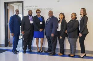 A group of seven professionals poses near a conference room sign, all dressed in business attire.