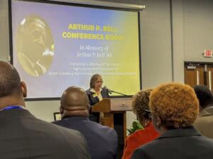 A speaker at a podium in a conference room honoring Arthur P. Bell, with attendees seated in the foreground.